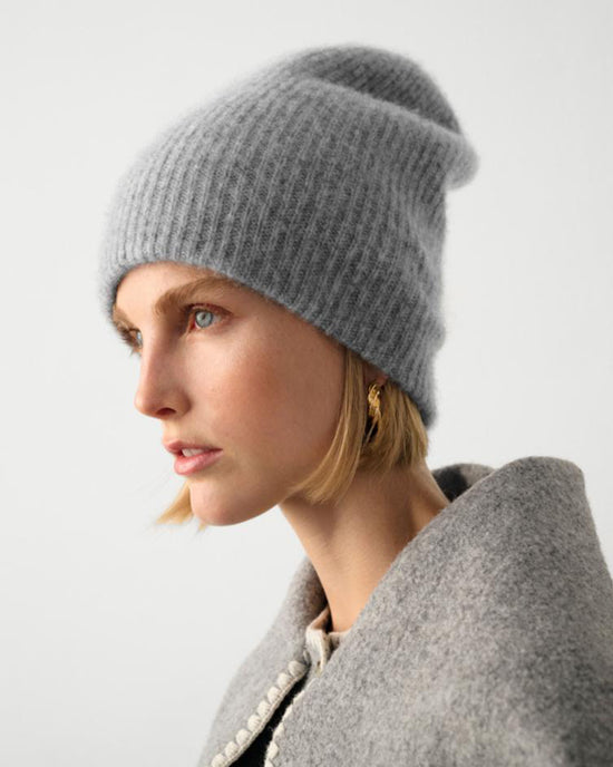 A woman with short blonde hair wears a White + Warren Cashmere Plush Rib Beanie in Grey Heather and a textured gray coat, gazing to the left against a plain light background.