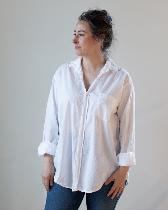 A woman in an oversized button-up, CP Shades Joss 1 Pocket in White Cotton Oxford shirt and jeans standing against a neutral background and looking to the side with a slight smile.
