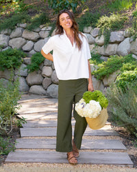 A woman smiles on a garden path, holding a basket of white and green flowers. She wears a white blouse, Frank & Eileen’s The Wexford Trouser in Hunter Green Linen, and sandals. Lush greenery and stone steps complete the scene.