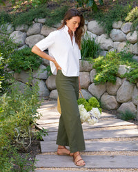 A woman wearing Frank & Eileens The Wexford Trouser in Hunter Green Linen and a white shirt walks along a wooden garden path, holding a basket of green and white flowers, surrounded by lush greenery and a stone wall.