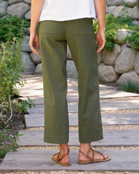A person stands on a wooden path outdoors, wearing Frank & Eileen’s The Wexford Trouser in Hunter Green Linen, a white shirt, and brown sandals. Stone wall and greenery are visible in the background. The photo is taken from behind.