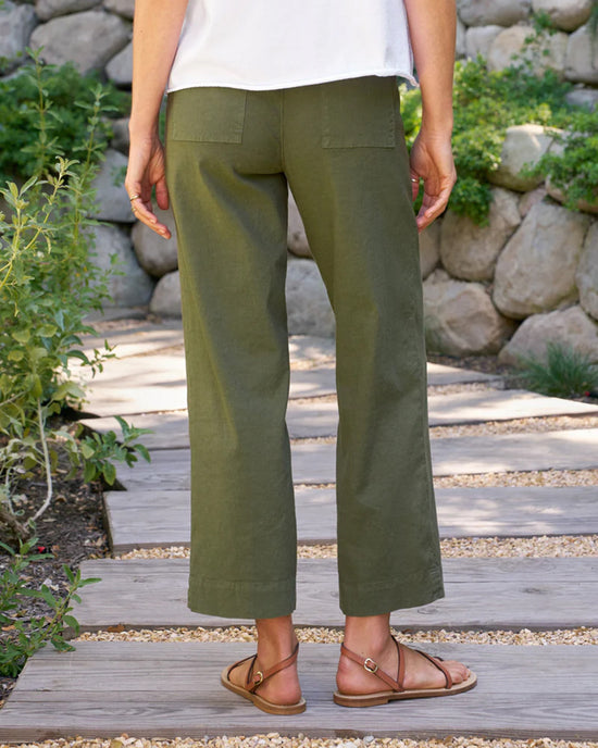 A person stands on a wooden path outdoors, wearing Frank & Eileen’s The Wexford Trouser in Hunter Green Linen, a white shirt, and brown sandals. Stone wall and greenery are visible in the background. The photo is taken from behind.