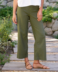 A person stands on a wooden path outdoors, wearing Frank & Eileen’s The Wexford Trouser in Hunter Green Linen with a white top and tan sandals. Relaxed among greenery and stone walls, they perfectly showcase this Italian Performance Linen style.