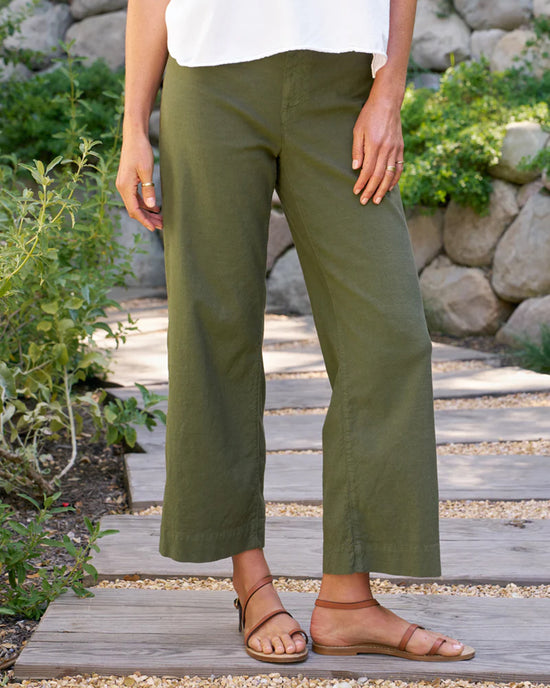 A person stands on a wooden path outdoors, wearing Frank & Eileen’s The Wexford Trouser in Hunter Green Linen with a white top and tan sandals. Relaxed among greenery and stone walls, they perfectly showcase this Italian Performance Linen style.