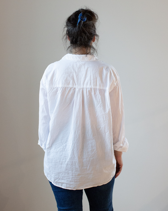 A person viewed from behind, wearing an oversized button-up Joss 1 Pocket in White Cotton Oxford CP Shades shirt and blue jeans, with hair tied up with a blue scrunchie.