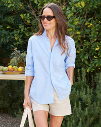 A woman with long brown hair, wearing sunglasses, the Eileen Relaxed Button-Up Shirt in Blue Casual Cotton by Frank & Eileen, and beige shorts, smiles while walking in a garden. She is holding a beige tote bag.