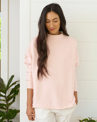 Woman in a pink Frank & Eileen Effie L/S Funnel Neck Capelet in Vintage Rose with closed eyes, standing in front of a white wall next to a green plant.