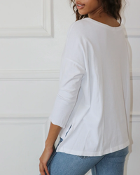 A person stands in front of a white wall, wearing a loose 100% cotton white Drop Shoulder Top in White by Felicite Apparel and blue jeans. The person is facing slightly away from the camera.