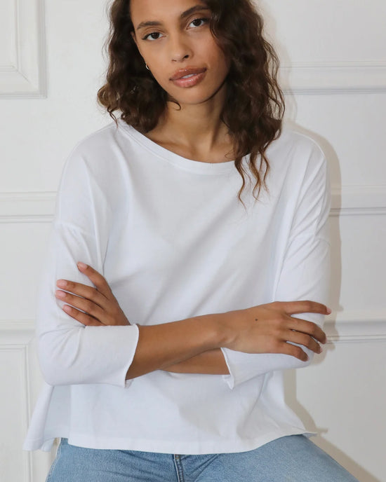 A woman in a white Drop Shoulder Top in White by Felicite Apparel and jeans stands with her arms crossed. She has long wavy hair and is looking directly at the camera against a white background.