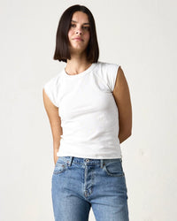 A person with straight, shoulder-length dark hair wears an NSF Chloe Fitted Slub Jersey Muscle Tee in Soft White and blue jeans, standing against a plain light-colored background.