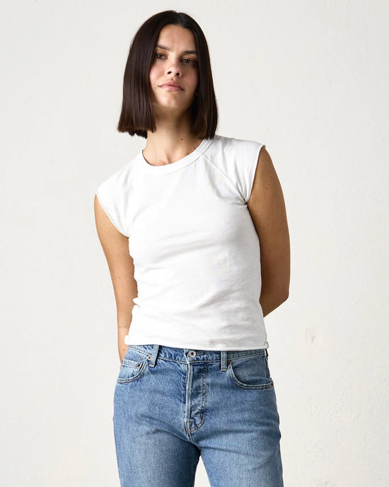 A person with straight, shoulder-length dark hair wears an NSF Chloe Fitted Slub Jersey Muscle Tee in Soft White and blue jeans, standing against a plain light-colored background.