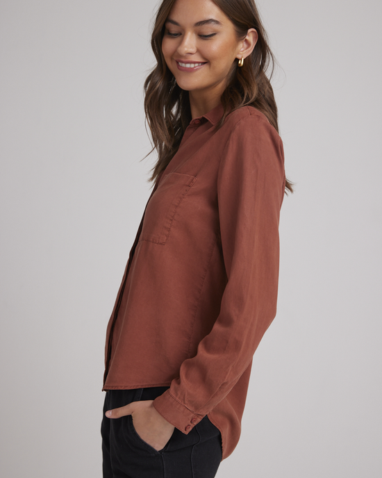 Woman smiling while posing in a casual Bella Dahl Two Pocket Classic Button Down in Autumn Amber with front button closure and dark pants.