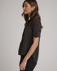 Side profile of a woman posing in a dark short-sleeved shirt layered over a white collar Bella Dahl Quartz Brown Tencel Split Back Button Down blouse.