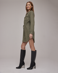 Woman smiling in a Bella Dahl Flap Pocket Shirt Dress in Herb Green made of TENCEL™ Lyocell, and black knee-high boots against a neutral background.