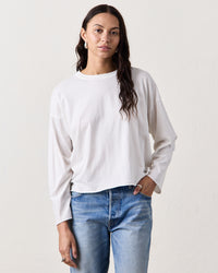 A person with long brown hair, dressed in the NSF Loffler Boxy L/S Crew in Chalk and blue jeans, stands against a plain light background.