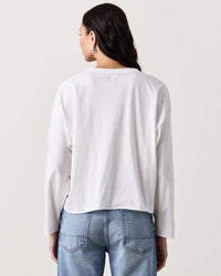 A woman stands with her back to the camera, wearing the NSF Loffler Boxy L/S Crew in Chalk and light blue jeans, set against a plain light background.