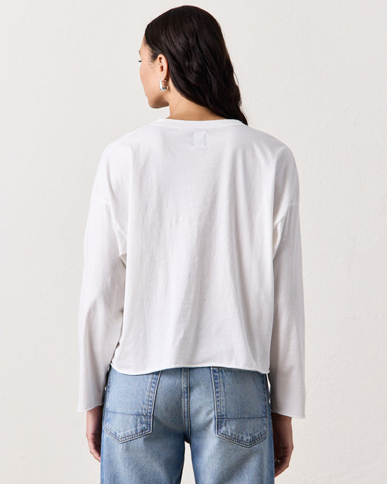 A woman stands with her back to the camera, wearing the NSF Loffler Boxy L/S Crew in Chalk and light blue jeans, set against a plain light background.