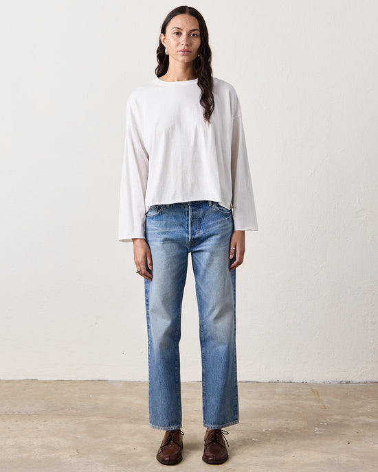 A woman stands indoors against a plain wall, wearing the NSF Loffler Boxy L/S Crew in Chalk, blue straight-leg jeans, and brown shoes.