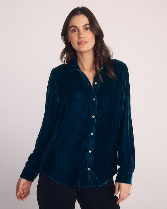 A woman with long dark hair wears a Bella Dahl Long Sleeve Classic Shirt in Harbor Teal and black pants, standing against a plain light background and looking at the camera with a neutral expression.