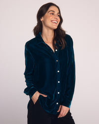 A woman with long brown hair smiles and looks to the side, wearing a Bella Dahl Long Sleeve Classic Shirt in Harbor Teal with black pants, standing against a plain light background.