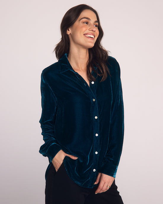A woman with long brown hair smiles and looks to the side, wearing a Bella Dahl Long Sleeve Classic Shirt in Harbor Teal with black pants, standing against a plain light background.