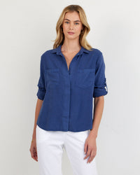A woman with blonde hair wears the Bella Dahl Split Back Button Down in Midnight Surf with rolled-up sleeves and white pants, standing against a plain light-colored background.