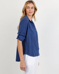 A woman with long blonde hair models the Bella Dahl Split Back Button Down in Midnight Surf, featuring rolled sleeves, paired with white pants, standing sideways and looking left against a plain white background.