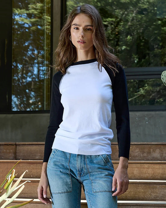 A woman with wavy brown hair wears the NSF Aria Fitted Baby Rib Raglan Tee in White/Black and blue jeans, standing outdoors by steps and a modern glass building, surrounded by greenery.