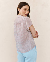 A woman with long dark hair wears the Bella Dahl Shirred Short Sleeve Shirt in Sunwashed Check and light blue pants, standing with her back to the camera and head turned to the side against a neutral background.