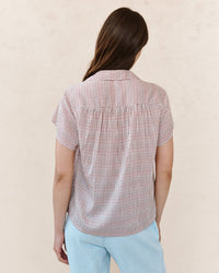 A person with long brown hair, facing away, wears the Bella Dahl Shirred Short Sleeve Shirt in Sunwashed Check and light blue pants against a plain light background.
