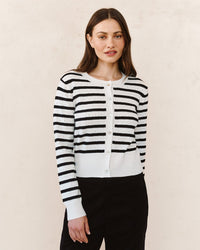 A woman with long brown hair and a neutral expression stands against a light background, wearing the Bella Dahl Fitted Cardigan in Cheri Stripe and fitted black pants.