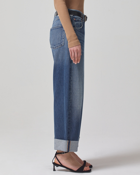 Side view of a person wearing cuffed Citizens of Humanity Ayla Baggy blue jeans made from organic cotton in Brielle and black strappy sandals, standing against a neutral background.