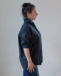 Profile view of a woman standing with her hands in her jeans pockets, wearing an oversized button-up Joss 1 Pocket in Ink Cotton Oxford from CP Shades.
