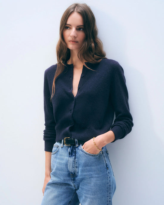 A woman with long, wavy brown hair wears a White + Warren Cashmere Crewneck Cardigan in Deep Navy and high-waisted blue jeans, standing against a plain light background with one hand in her pocket.