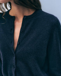 A woman with wavy brown hair wears the White + Warren Cashmere Crewneck Cardigan in Deep Navy, top buttons undone to reveal her collarbone. The background is a plain light gray wall.