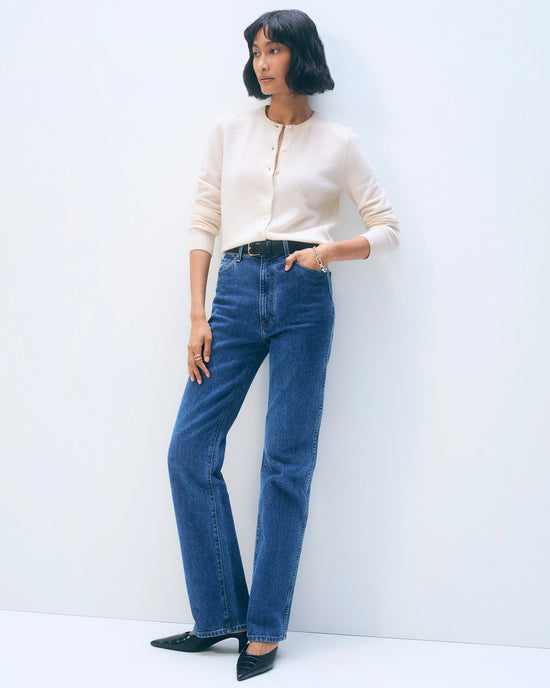 A woman stands against a light background wearing the White + Warren Cashmere Crewneck Cardigan in Soft White, paired with high-waisted blue jeans, a black belt, and black pointed-toe shoes. One hand is in her pocket as she looks to the side.