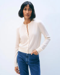 A woman with short dark hair wears a White + Warren Cashmere Crewneck Cardigan in Soft White and blue jeans, standing against a plain light background with one hand in her pocket.