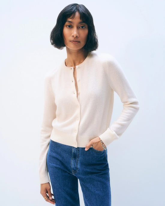 A woman with short dark hair wears a White + Warren Cashmere Crewneck Cardigan in Soft White and blue jeans, standing against a plain light background with one hand in her pocket.