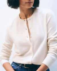 Someone wears the White + Warren Cashmere Crewneck Cardigan in Soft White, with a few buttons undone, styled with blue jeans and a black belt. A plain background and partially cropped face give the outfit a relaxed, casual vibe.