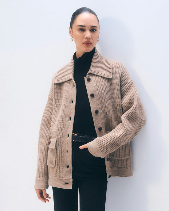 A woman with slicked-back hair wears the White + Warren Merino Fisherman Jacket in Macadamia over a black turtleneck and pants, standing against a light background with one hand in her pocket and a neutral expression.