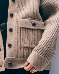 Close-up of a person wearing the White + Warren Merino Fisherman Jacket in Macadamia, crafted from Scottish merino wool with large dark buttons and a front pocket. Their gold-ringed hand rests near the pocket.
