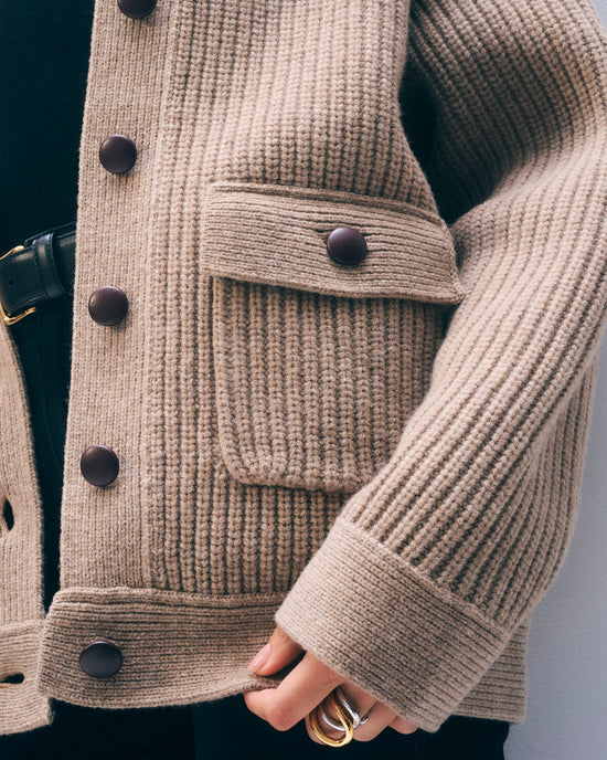 Close-up of a person wearing the White + Warren Merino Fisherman Jacket in Macadamia, crafted from Scottish merino wool with large dark buttons and a front pocket. Their gold-ringed hand rests near the pocket.