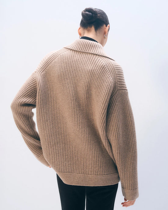 A person with dark hair in a neat bun is shown from behind, wearing the White + Warren Merino Fisherman Jacket in Macadamia with black pants against a light, plain background.