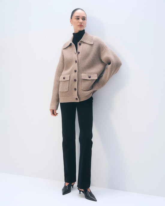A woman stands against a white background wearing the White + Warren Merino Fisherman Jacket in Macadamia, featuring large pockets, paired with a black turtleneck, slim black pants, and pointed black heels.