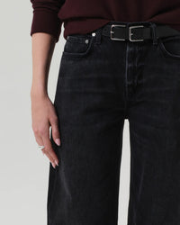 Wearing Citizens of Humanity's Miro Relaxed Jean in Raine, styled with a black belt and dark maroon sweater. Their left hand, adorned with light pink nails and rings, rests by their side against a plain light background.