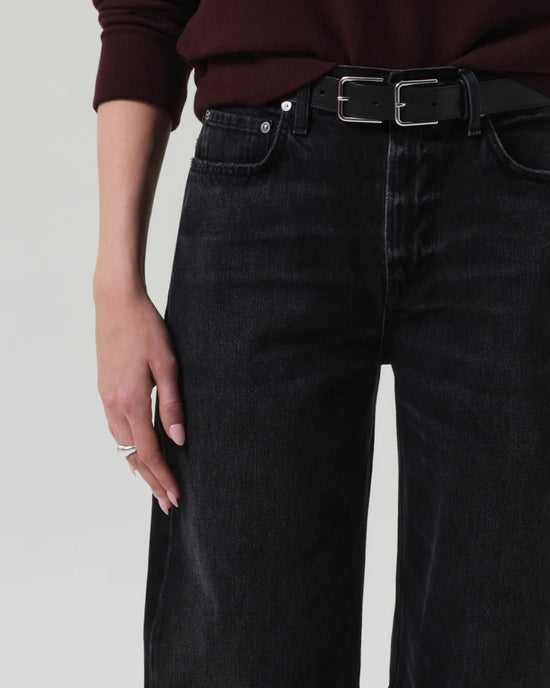 Wearing Citizens of Humanity's Miro Relaxed Jean in Raine, styled with a black belt and dark maroon sweater. Their left hand, adorned with light pink nails and rings, rests by their side against a plain light background.