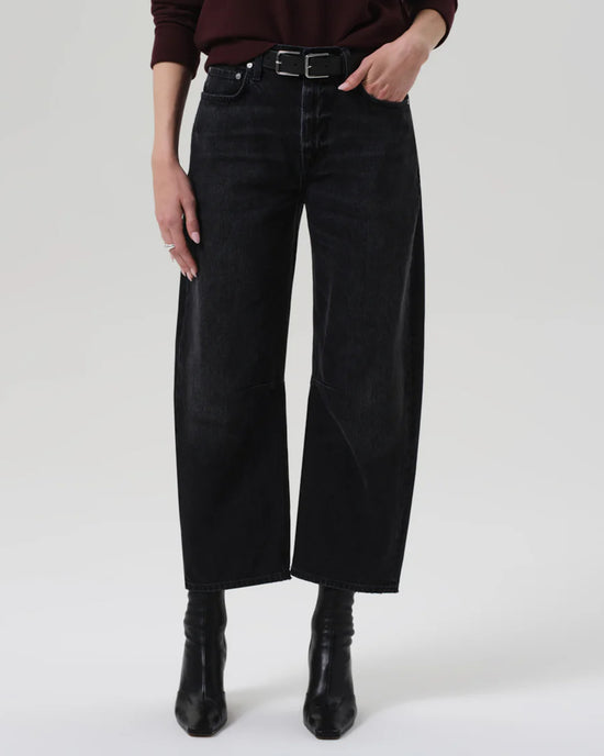 A person wearing Citizens of Humanity’s Miro Relaxed Jean in Raine, styled with a belt, black boots, and a dark long-sleeve top, stands against a white background. Only the lower torso and legs are visible.
