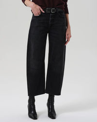 A person wears Citizens of Humanity’s Miro Relaxed Jean in Raine—high-rise, washed black denim made from regenerative cotton—styled with a black belt, dark long-sleeve top, and black heeled boots against a light background.