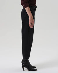A person, shown from mid-torso down, stands in profile wearing Citizens of Humanity’s Miro Relaxed Jean in Raine—high-rise barrel-leg jeans made from regenerative cotton in washed black denim—paired with a dark long-sleeve top and black pointed-toe heeled boots.