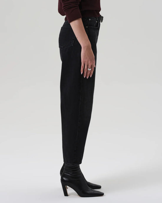 A person, shown from mid-torso down, stands in profile wearing Citizens of Humanity’s Miro Relaxed Jean in Raine—high-rise barrel-leg jeans made from regenerative cotton in washed black denim—paired with a dark long-sleeve top and black pointed-toe heeled boots.